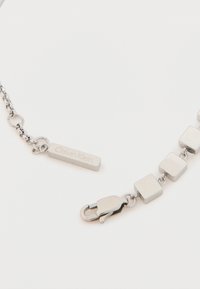 Sidabrinės spalvos vėrinys su kvadratiniais akmenėliais, su naginėmis užsegimu ir stačiakampiu žymekliu, kuriame išgraviruotas "Calvin Klein". Lygus metalinis paviršius.