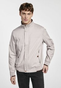 Jasnoszara kurtka na zamek z wysokim kołnierzem, z bocznymi kieszeniami, elastycznymi mankietami i brązowymi akcentami. Wykonana z gładkiego, lekkiego materiału.