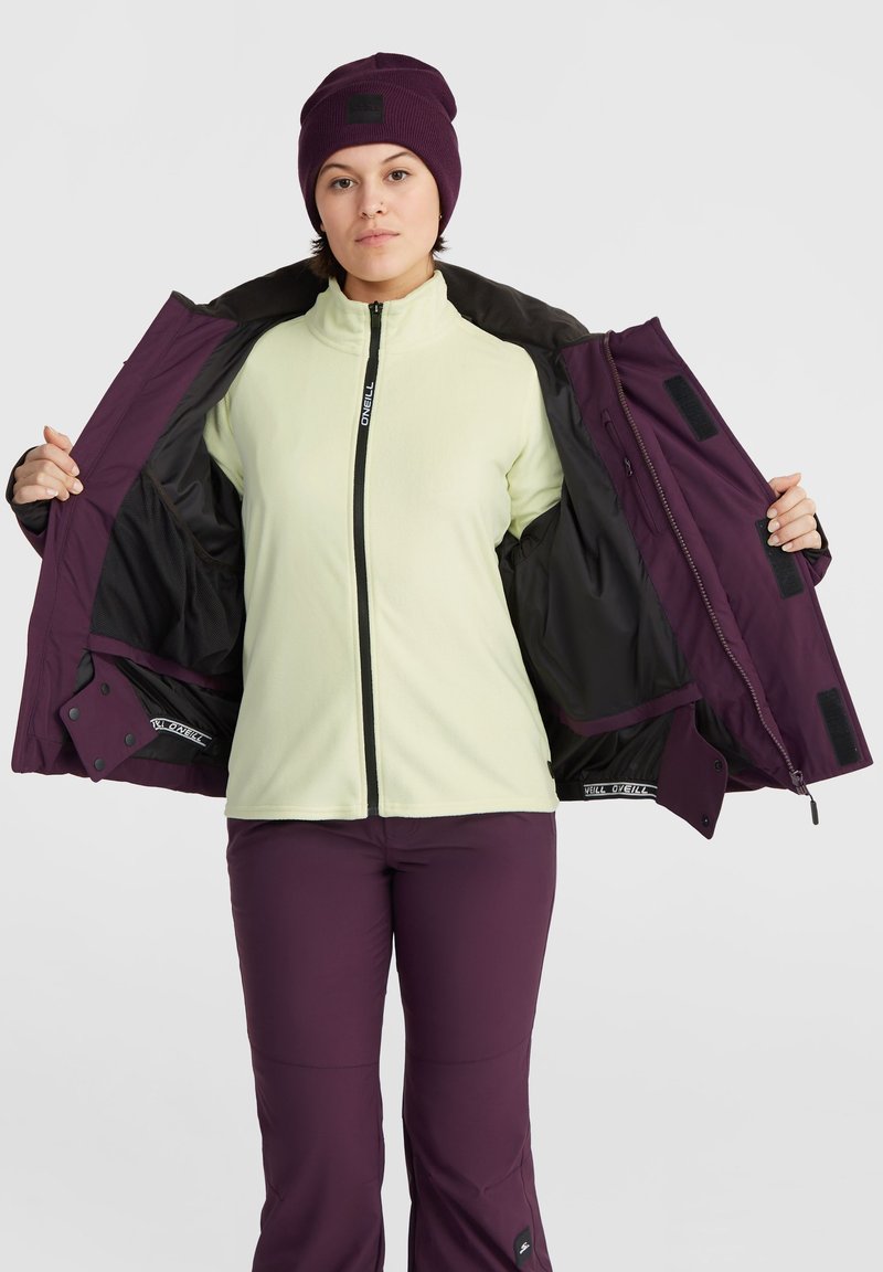 Lila Jacke mit schwarzer Fütterung, Reißverschlussverschluss und Klettverschluss-Akzenten, getragen über einem hellgrünen Fleece-Oberteil und passenden lila Hosen.