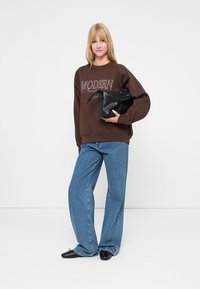 Sweatshirt marron avec un graphique "MODERN", associé à un jean large bleu clair. Tenant un petit sac noir texturé. Chaussures plates noires.