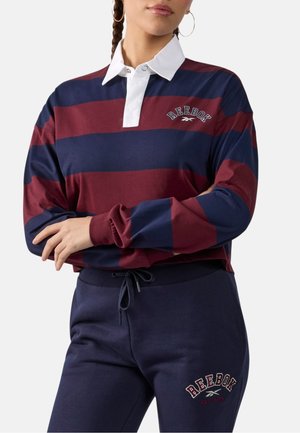 Persona que lleva una camiseta de rugby Reebok a rayas burdeos y azul marino con cuello blanco y pantalones deportivos Reebok azul marino con cintura de cordón.