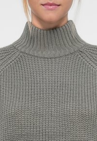 Lys grå strikket genser med høy, ribbet krage og et strukturert mønster. Stoffet har et mykt, tykt utseende.