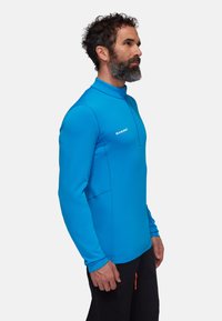 Blå långärmad tröja med halvmatchande krage, tillverkad av ett mjukt, stretchigt material. Har en logotyp på bröstet och platta sömmar för komfort.