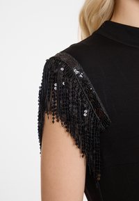 Zwarte jurk met ingewikkelde franjes van kralen op de schouders, die textuur en visuele interesse toevoegen aan het strakke ontwerp.