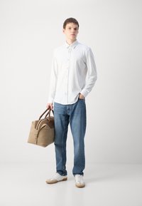 Camisa branca de botões com padrão subtil, jeans azul claro e ténis cinzentos. A segurar uma mochila de duffel castanha e bege com alças duplas.