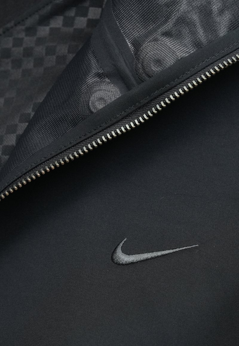 Black fabric with a silver Nike swoosh logo and a partially unzipped black zipper revealing patterned inner lining.