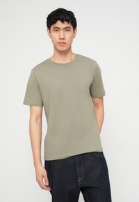 Νεαρός με κοντά μαύρα μαλλιά που φοράει απλό πουκάμισο ελιάς και σκούρο μπλε τζιν, στέκεται μπροστά σε ανοιχτό φόντο.