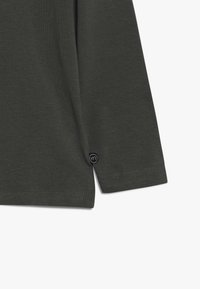 Dunkelgrünes Langarmshirt mit gerippter Textur, seitlichen Schlitzen und einem kleinen schwarzen Knopf-Detail am Saum. Glattes Baumwollmaterial.