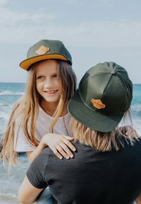 Due cappelli verdi e gialli con un logo marrone, indossati da un bambino e un adulto in spiaggia, con onde blu dell'oceano sullo sfondo.