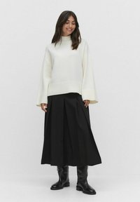 Kvinde iført en hvid oversized sweater, sort plisseret midi-nederdel og sorte knæhøje støvler, der står imod en ensfarvet lys baggrund.