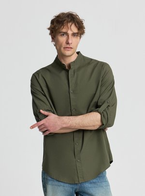 Mężczyzna z kręconymi włosami ubrany w oliwkowozieloną koszulę na guziki z podwiniętymi rękawami i jasnoniebieskie dżinsy, pozujący ze skrzyżowanymi ramionami.