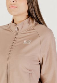 Chaqueta de cremallera en color beige claro que presenta un cuello alto, una textura suave y el logo de EA7 en el pecho. Diseño minimalista con costuras sutiles.