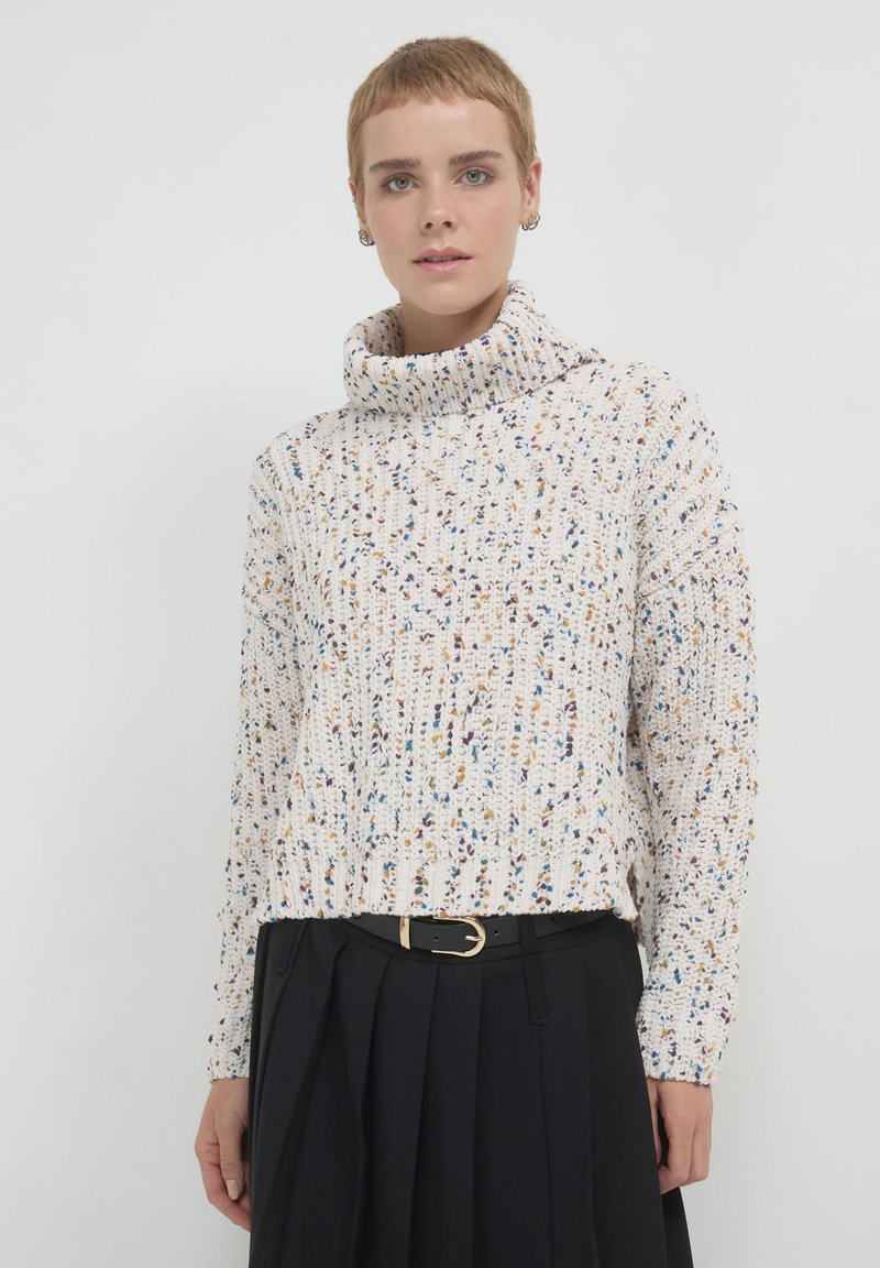 Pull à col roulé de couleur claire avec un motif texturé, présentant des éclats de bleu, jaune et violet. Assorti avec une jupe plissée noire.