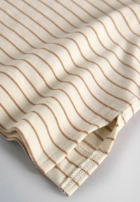 Närbild av beige tyg med tunna horisontella bruna ränder och en liten sidospalt vid fållen som visar tygstruktur och sömdetaljer.