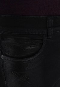 Jean en denim noir avec une texture lisse, présentant une poche avant, un bouton en métal et des coutures visibles. Le matériau a un léger éclat.