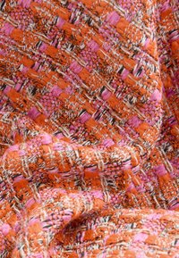 Textiel met een oranje en roze geweven patroon, met een gestructureerd oppervlak en zichtbare vezels, wat een dichte, gelaagde uitstraling creëert.