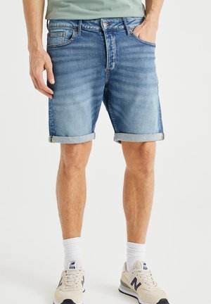 Mężczyzna noszący podwinięte niebieskie szorty dżinsowe, białe skarpety i beżowe buty New Balance, stojący z jedną ręką w kieszeni.