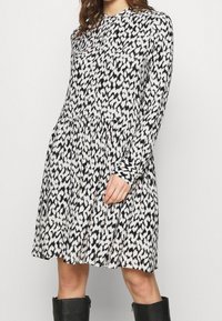 Långärmad klänning i svart och vit djurtryck, med hög krage, plisserad midja och en avslappnad silhuett. Materialet verkar mjukt.