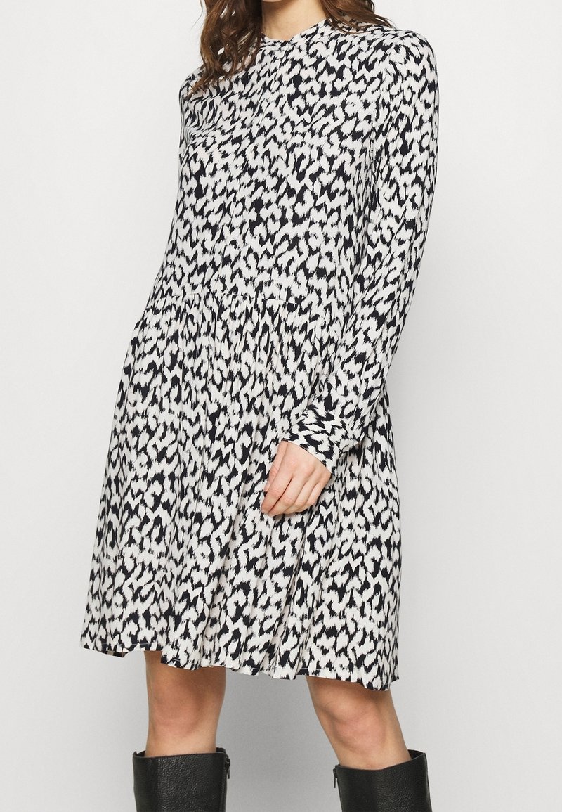 Långärmad klänning i svart och vit djurtryck, med hög krage, plisserad midja och en avslappnad silhuett. Materialet verkar mjukt.