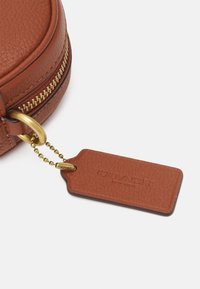 Borsa rotonda in pelle marrone con superficie texturizzata, zip dorata e un'etichetta abbinata attaccata da una catena, stampata con "COACH" e "NEW YORK."