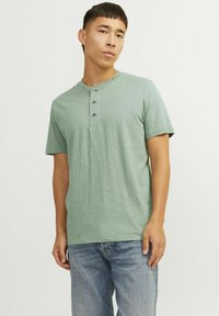 Camiseta henley de manga corta en verde apagado, hecha de tejido ligero, presenta una tapeta de tres botones y un escote redondo. Se lleva con vaqueros azules.