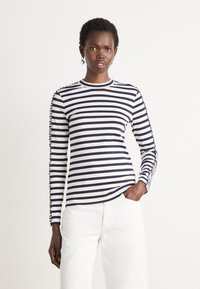 Maglietta a maniche lunghe a righe in cotone blu navy e bianco, con scollatura rotonda e dettagli intricati lungo le maniche, abbinata a pantaloni bianchi.