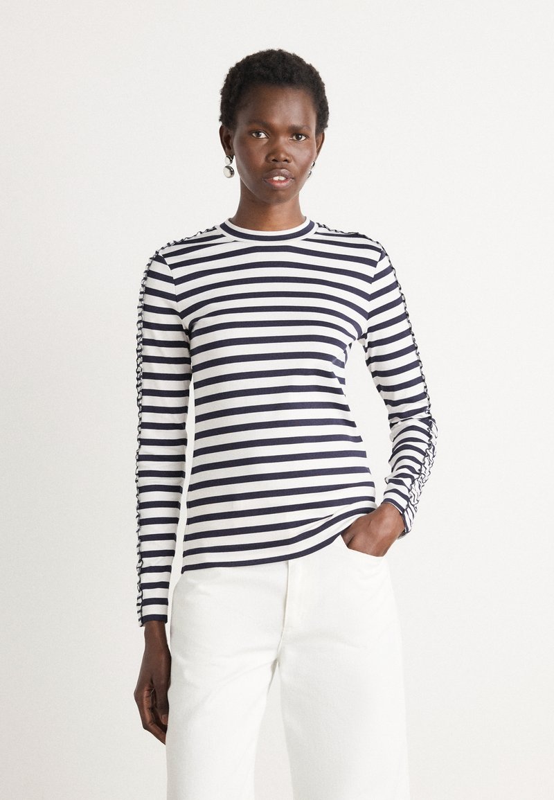 Maglietta a maniche lunghe a righe in cotone blu navy e bianco, con scollatura rotonda e dettagli intricati lungo le maniche, abbinata a pantaloni bianchi.