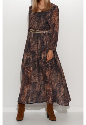 Femme portant une longue robe imprimée serpent marron et noire avec une ceinture chaîne dorée et des bottines en daim marron.
