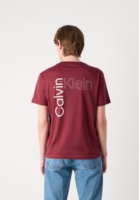 Bordová bavlnená tričko s bielym logom na chrbte, s krátkymi rukávmi a okrúhlym výstrihom, v kombinácii s svetlomodrými džínsy.