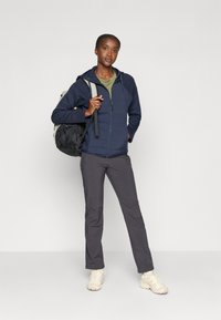 Navy zip-up kapuce, ar teksturētu virsmu, kombinēts ar pelēkiem biksēm. Modelis tur melnu mugursomu un valkā bežīgas sporta kurpes.