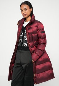Burgundská péřová bunda s vysokým límcem, lesklou texturou, zlatými doplňky a bočním logem, nošená přes černý vzorovaný svetr.