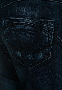 Mörkblå denimjeans som har en bakfickor med ett böjt lock, synliga sömmar och en lätt sliten yta.