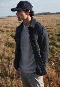 Chaqueta de cuadros grises tejida sobre un suéter claro. Una gorra oscura y pantalones beiges completan el conjunto, con un fondo de campo de hierba.