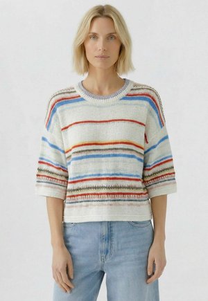 Blonde Frau trägt einen weißen Strickpullover mit mehrfarbigen horizontalen Streifen und hellblaue Jeans, steht vor einem schlichten Hintergrund.