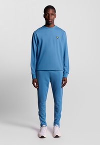 Felpa azzurra chiara e pantaloni jogger coordinati, realizzati in morbido tessuto, con un piccolo logo giallo sul petto. Sneakers bianche completano l'outfit.