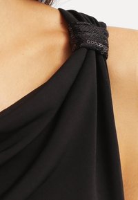 Svart tygklänning med en draperad design och en texturerad paljettdetalj på axeln som f örstärker den övergripande formen och visuella detaljer.