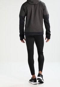 Atletisk jacka i mörkgrått tyg med huva och svarta detaljer, tillsammans med åtsittande svarta leggings och svarta träningsskor.