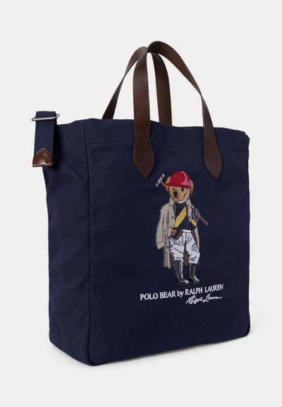 Marineblå handlenett med brune lærhåndtak, med en brodert bjørn i rytterantrekk og rød hjelm som holder en polokølle.