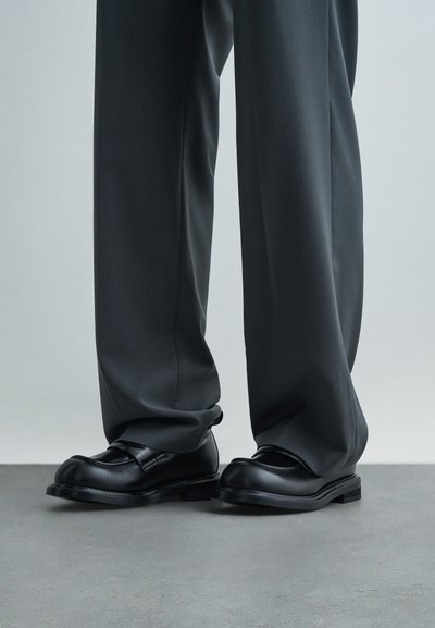 Cilvēks valkā tumši pelēkus brīvus bikšu un melnas ādas kurpes, stāvot pelēkā grīdā uz gaiša fona.