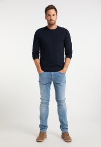 Tengerészkék kötött pulcsi, karcsú szabású kék farmer és bézs velúr cipők. A modell kezeit a zsebében tartva áll egy sima fehér háttér előtt.