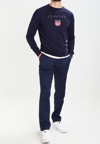 Navy sweatshirt met wit logo en schildembleem, gecombineerd met navy broek. Het model draagt witte sneakers en staat op een neutrale achtergrond.