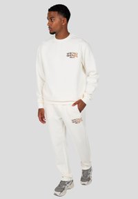 Conjunto de sudadera blanca con sudadera de cuello redondo y pantalones a juego. Lleva el logo "WRONG FRIENDS" en marrón. Tejido suave y texturizado. Zapatillas grises.