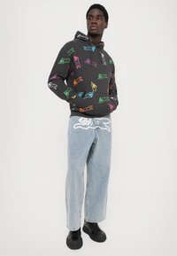 O hoodie apresenta um tecido preto com contornos de animais coloridos, combinado com calças jeans largas de um azul claro e sapatilhas pretas.