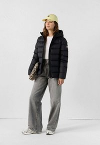 Sort pufferjakke med hætte, hvid t-shirt, grå brede jeans, taske med leopardprint og lys gul kasket. Sølv sneakers fuldender looket.