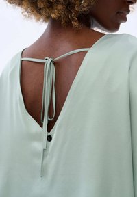 Zielona bluzka bez pleców z głębokim dekoltem w kształcie V i regulowanymi trokami. Gładki, lekki materiał z małym czarnym detalem guzika.