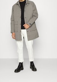 Abrigo de cuadros en beige y negro con un corte relajado, que presenta un cuello, cierre de botones y grandes bolsillos frontales. Llevado sobre una blusa negra y pantalones blancos.