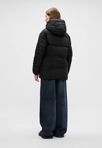 Mustat puffatakki, jossa on huppu ja tikattu muotoilu. Yhdistetty leveälahkeisiin haalistuneisiin sinisiin farkkuihin, katsottuna takaa.