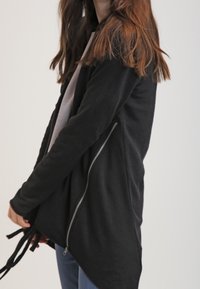Černá zipová bunda s drapovaným designem, která má diagonální zip a detaily se stahovací šňůrkou, vyrobená z měkkého textilního materiálu.