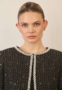 Chaqueta de tweed negra con cuello redondo adornado con perlas y embellishments metálicos, diseño estructurado y detalles de costura visibles.