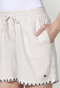 Shorts en lin beige clair avec une ceinture élastique, un cordon de serrage et des poches latérales. Ornés d'une broderie de feuilles noires le long de l'ourlet.